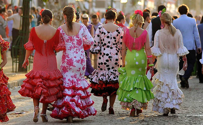 Women in traditional Sevillanas dance dresses walking down a crowded cobblestone street.